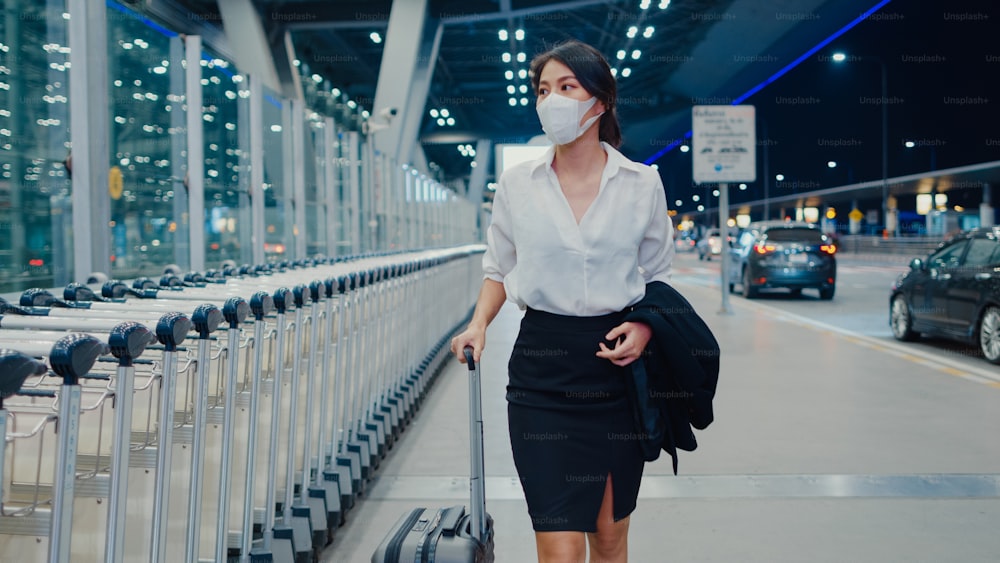 아시아 비즈니스 소녀 도착 목적지는 국내 공항의 대기 차량 터미널 밖에서 드래그 수하물 산책으로 얼굴 마스크를 착용합니다. 비즈니스 통근 covid 전염병, 비즈니스 여행 사회적 거리두기 개념.