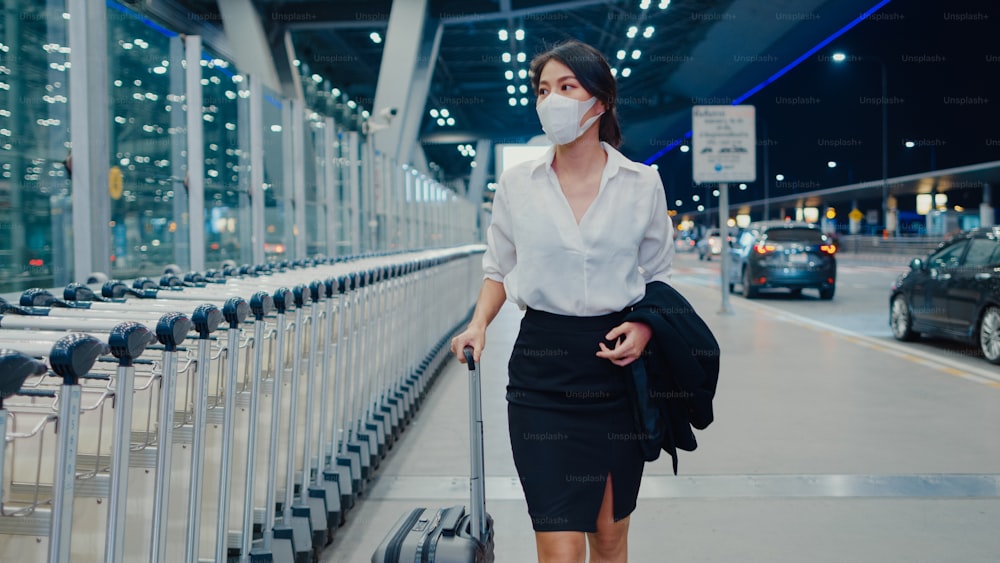 Asian business girl arrive destination wear face mask with drag luggage walk outside wait car terminal at domestic airport. Business commuter covid pandemic, Business travel social distancing concept.
