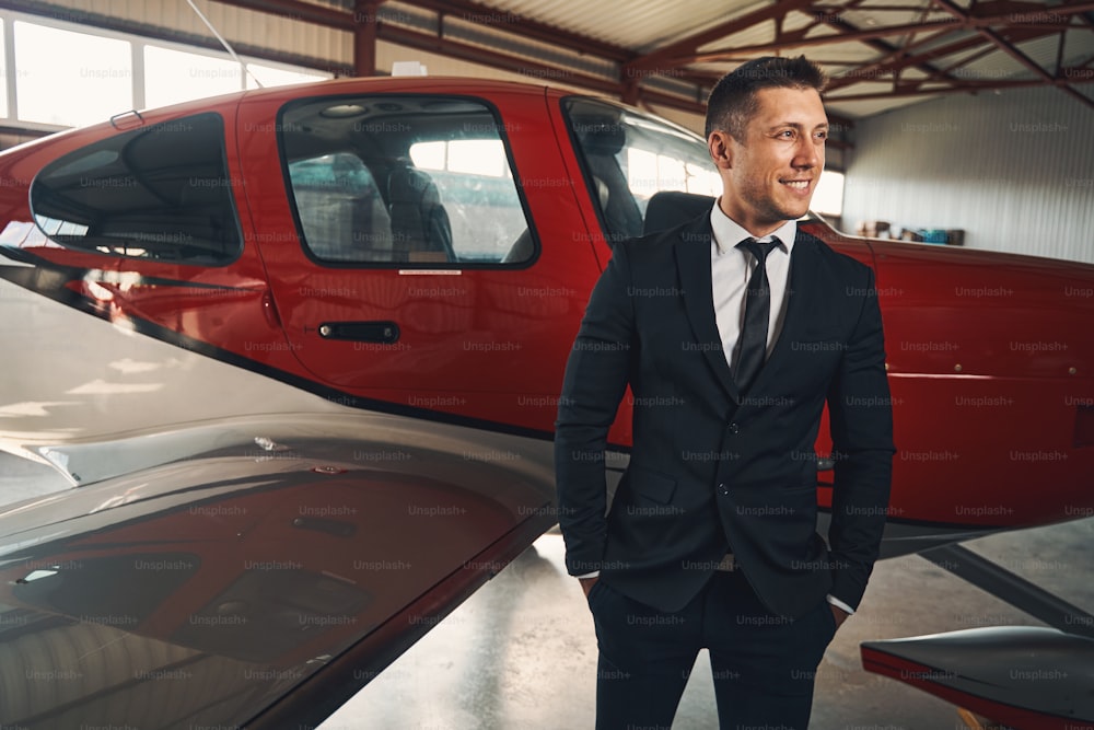 Handsome businessman in suit keeping hands in pockets and smiling while standing near aircraft