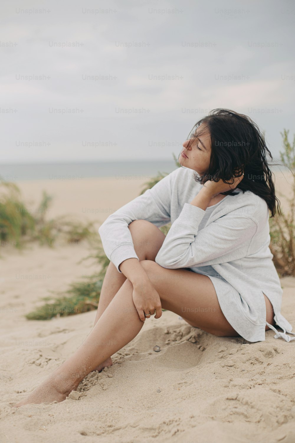 Beautiful woman with windy hair sitting on sandy beach on background of green grass and sea, calm tranquil moment. Stylish young female in sweater relaxing on coast