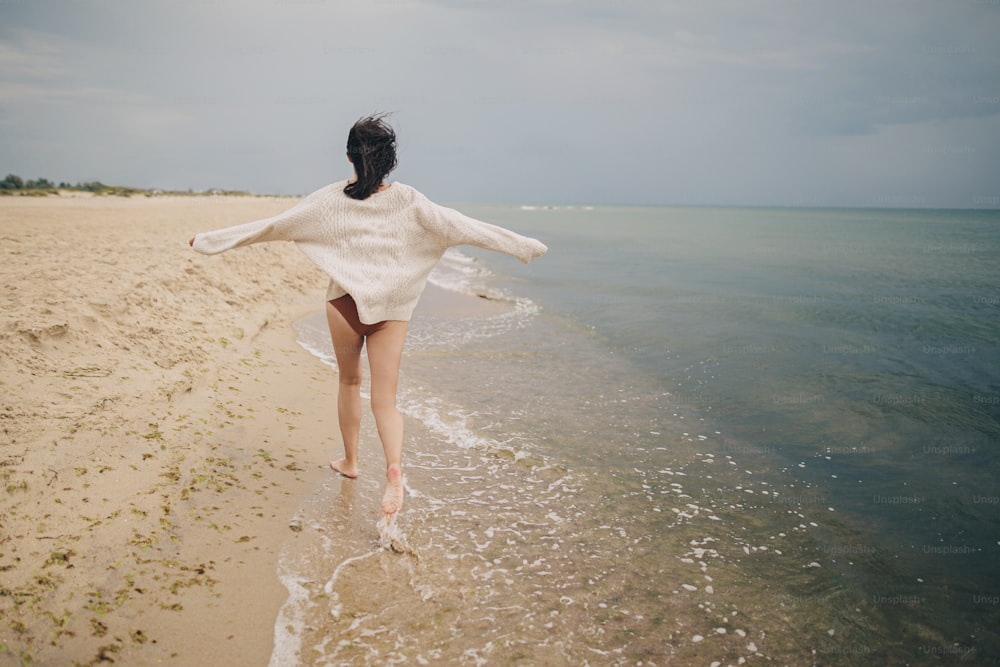 ニットセーターを着て、冷たい海の砂浜を走っている風の強い髪の屈託のない美しい女性。スタイリッシュな若い幸せな女性がリラックスして海岸で休暇を楽しんでいます。背面図