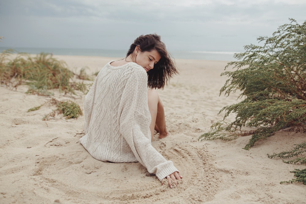Beautiful woman with windy hair sitting on sandy beach with green grass, carefree tranquil moment. Stylish young sexy female in knitted sweater holding sand and relaxing on coast