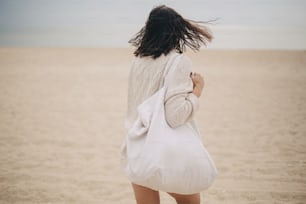 Beautiful stylish woman with windy hair and tote bag walking on sandy beach to sea, carefree moment. Stylish young female in knitted sweater enjoying vacation and relaxing on coast