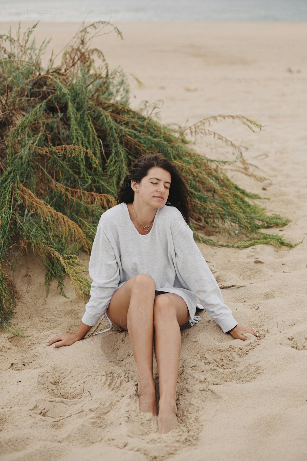 Beautiful woman with windy hair and in sweater sitting on sandy beach on background of green grass and sea, calm tranquil moment. Stylish young female relaxing on coast. Vacation mood