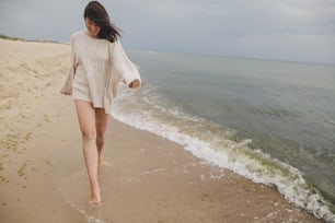 Carefree beautiful woman with windy hair running on sandy beach at cold sea waves, having fun. Stylish young happy female in knitted sweater relaxing and enjoying vacation on coast