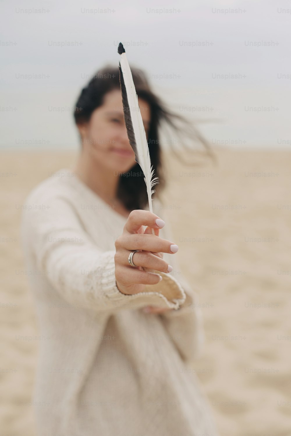 Beautiful woman with windy hair holding feather on background of sandy beach and sea, tranquil moment. Stylish young boho female in sweater holding bird feather on coast. Free and wild concept