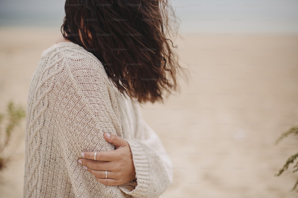 Beautiful woman with windy hair sitting on sandy beach on background of sea, calm tranquil moment. Closeup of stylish young female hand on knitted sweater on cold coast