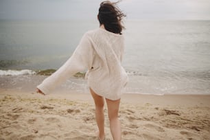 Carefree beautiful woman in knitted sweater and with windy hair running on sandy beach at cold sea, having fun. Stylish young happy female relaxing and enjoying vacation on coast. Back view