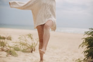 Sand on background of beautiful woman legs barefoot walking on sandy beach and having fun, close up. Carefree vacation mood. Stylish young female in knitted sweater relaxing on coast