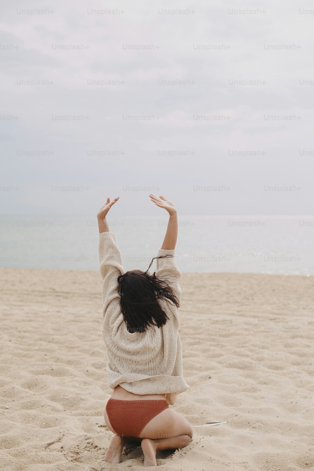 Bella donna felice con i capelli ventosi seduta sulla spiaggia sabbiosa in mare, momento spensierato. Giovane donna elegante in maglione lavorato a maglia e bikini che si gode le vacanze e si rilassa. Vista posteriore. Immagine verticale