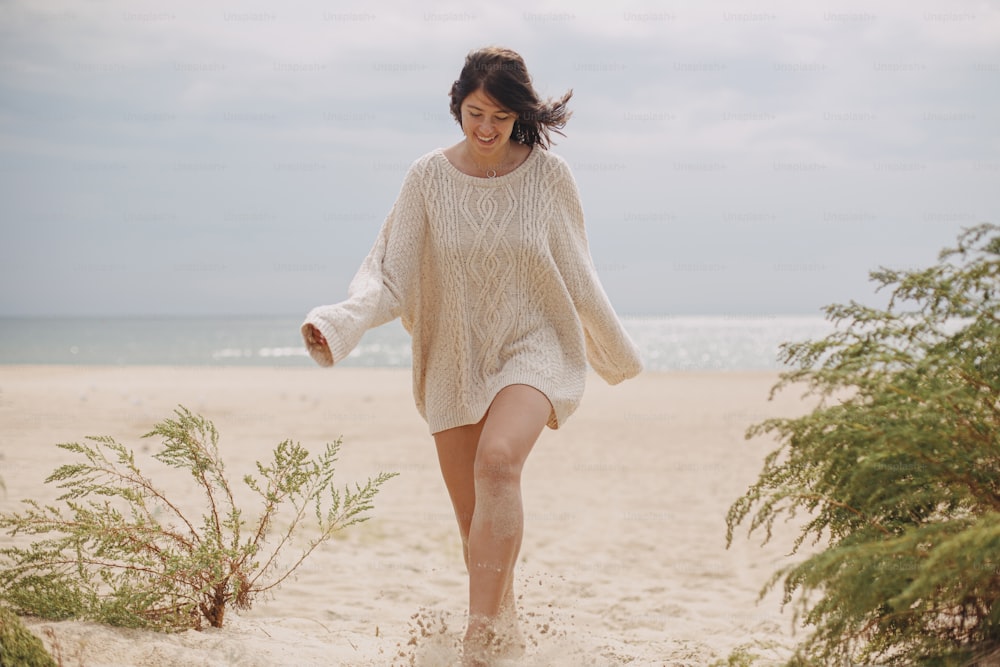 Mulher feliz bonita com cabelo ventoso andando na praia de areia no fundo da grama verde e do mar, momento calmo e tranquilo. Fêmea jovem elegante no suéter de malha bege que relaxa na costa fria