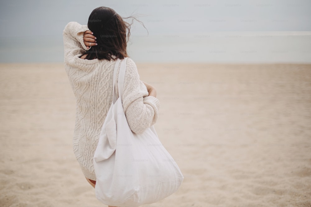 Beautiful stylish woman with windy hair and tote bag walking on sandy beach to sea, carefree moment. Stylish young female in knitted sweater enjoying vacation and relaxing on coast. Copy space