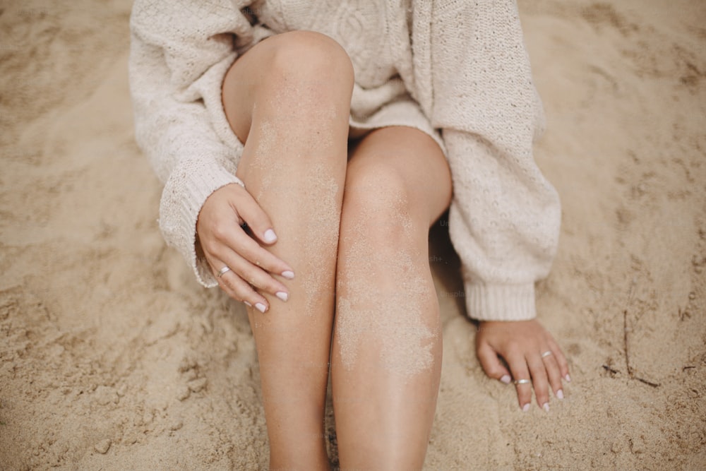 Hermosa mujer con piernas bronceadas en arena sentada en la playa de arena con hierba verde, de cerca. Momento tranquilo y sin preocupaciones. Mujer sexy joven con estilo en suéter que se relaja en la costa