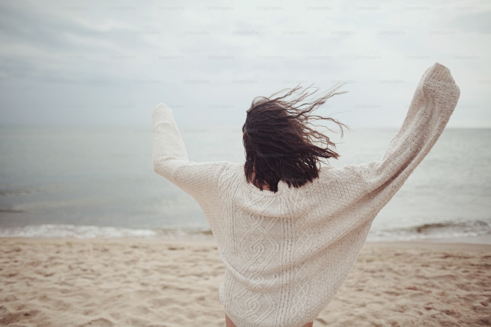 ニットセーターを着て、冷たい海の砂浜を走っている風の強い髪の屈託のない美しい女性。スタイリッシュな若い幸せな女性がリラックスして海岸で休暇を楽しんでいます。背面図