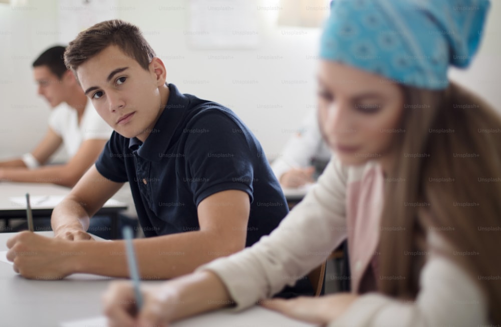 Teenagers students sitting in the classroom working exam. Male looking at camera. Focus is on background.