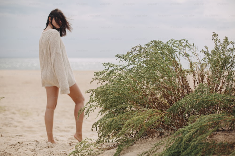 Hierba verde en la playa de arena en el fondo de la silueta borrosa de la mujer hermosa con el pelo ventoso, momento tranquilo y tranquilo. Elegante joven femenina en suéter de punto beige relajándose en la costa