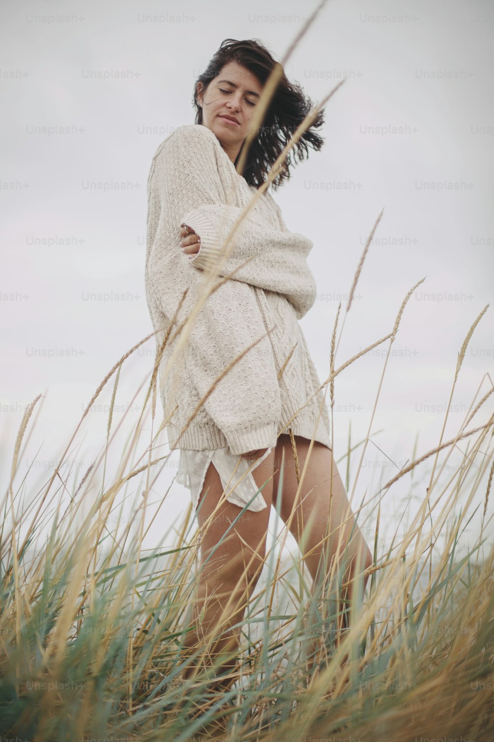 니트 스웨터를 입고 바람이 부는 머리를 한 아름다운 세련된 여성이 바다의 야생 잔디 사이에서 포즈를 취하고 있습니다. 진정한 평온한 순간. 바람이 부는 해안에 서 있는 세련된 젊은 여성. 평온. 세로