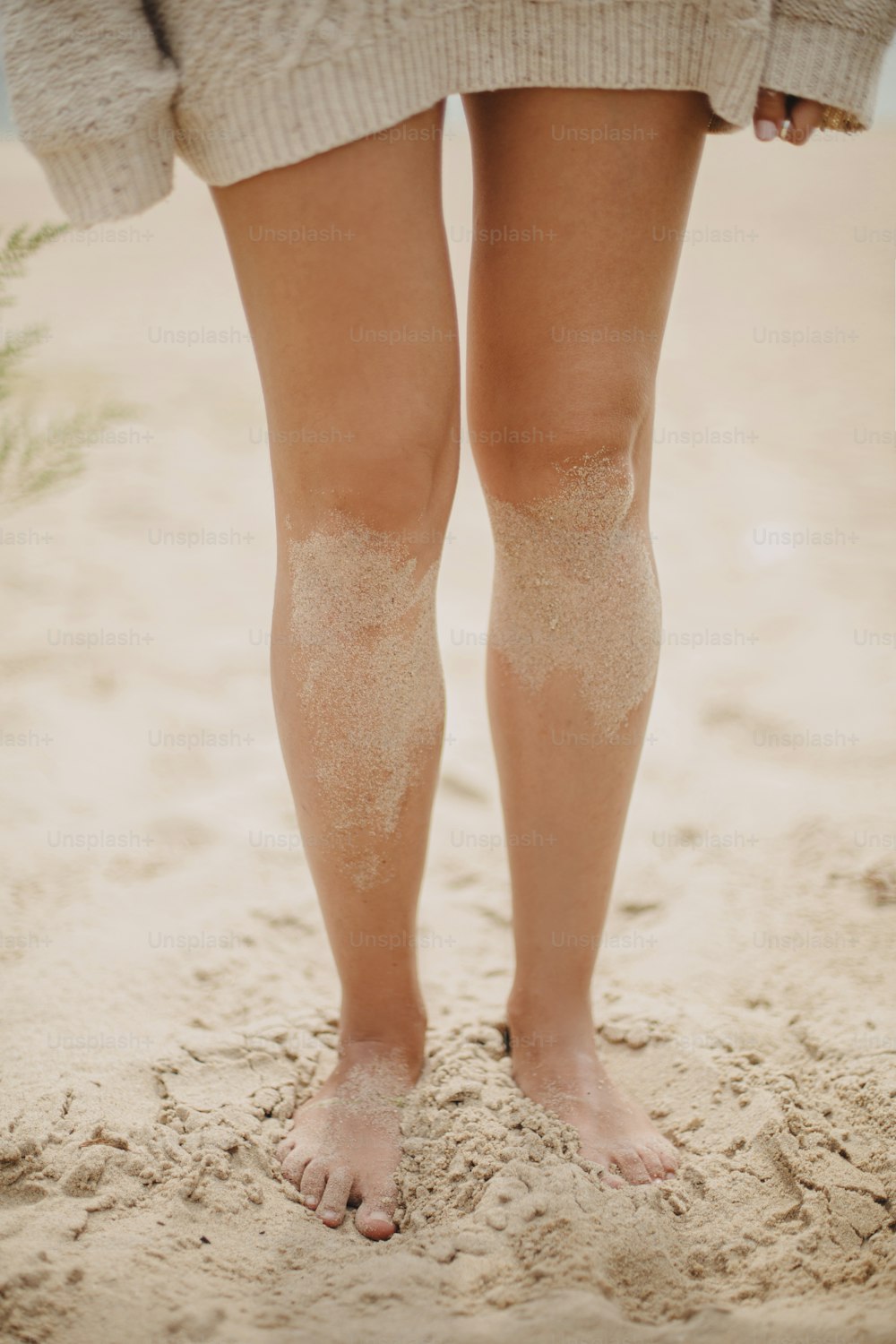 Beautiful woman legs barefoot with sand standing on sandy beach, carefree vacation mood. Stylish young female in knitted sweater relaxing on coast, cropped view. Vertical image