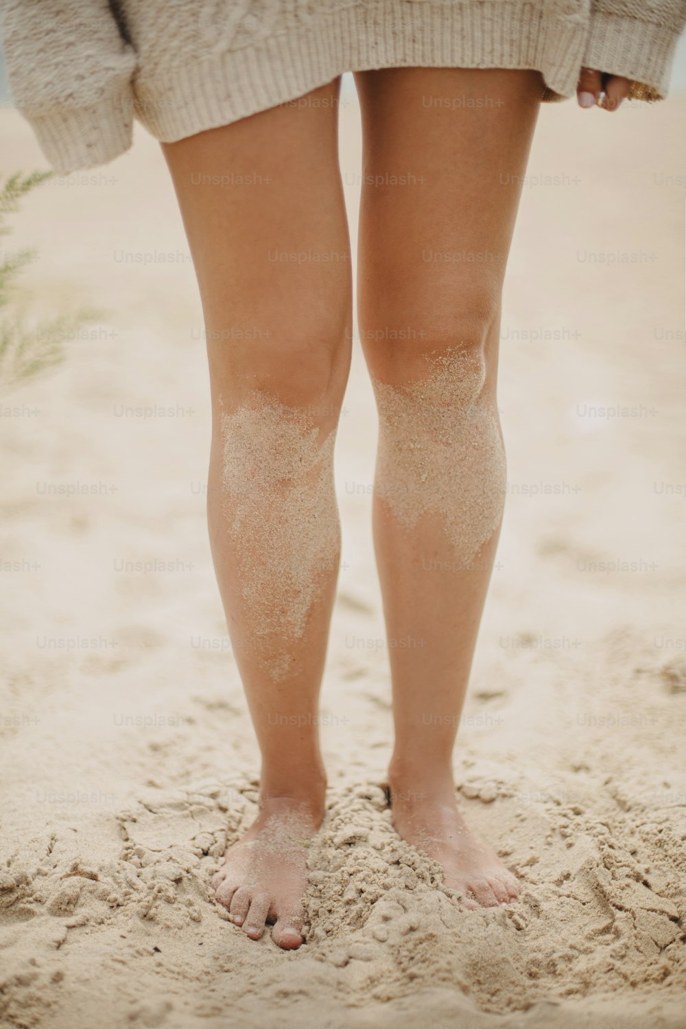 모래 해변에 서 있는 모래와 함께 맨발로 아름다운 여자 다리, 평온한 휴가 분위기. 니트 스웨터를 입은 세련된 젊은 여성이 해안에서 휴식을 취하고 있다. 세로 이미지