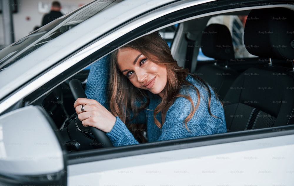 Donna positiva in camicia blu si siede all'interno di una nuova auto nuova di zecca. Nel salone dell'automobile o nell'aeroporto.