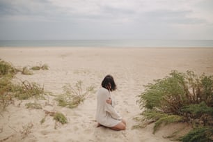 Bella donna con i capelli ventosi seduta sulla spiaggia sabbiosa sullo sfondo dell'erba verde e del mare, momento tranquillo calmo. Giovane donna elegante in maglione lavorato a maglia che si rilassa sulla costa fredda