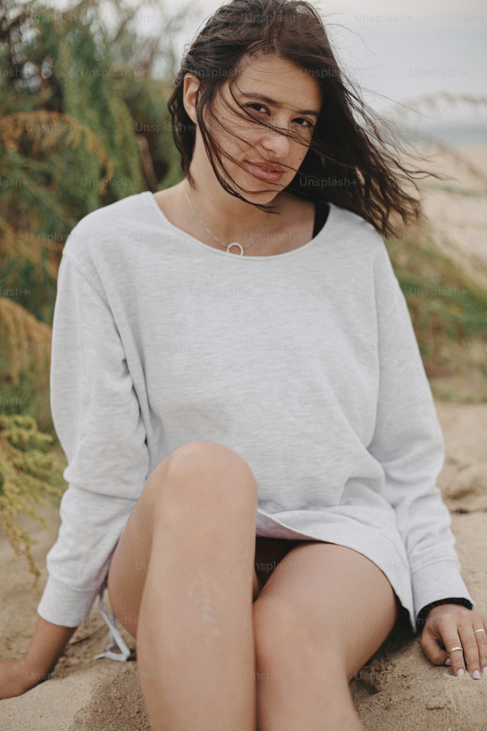 Mulher despreocupada bonita com cabelo ventoso e em suéter sentado na praia de areia no fundo da grama verde e do mar, momento tranquilo. Jovem fêmea elegante relaxando na costa. Clima de férias