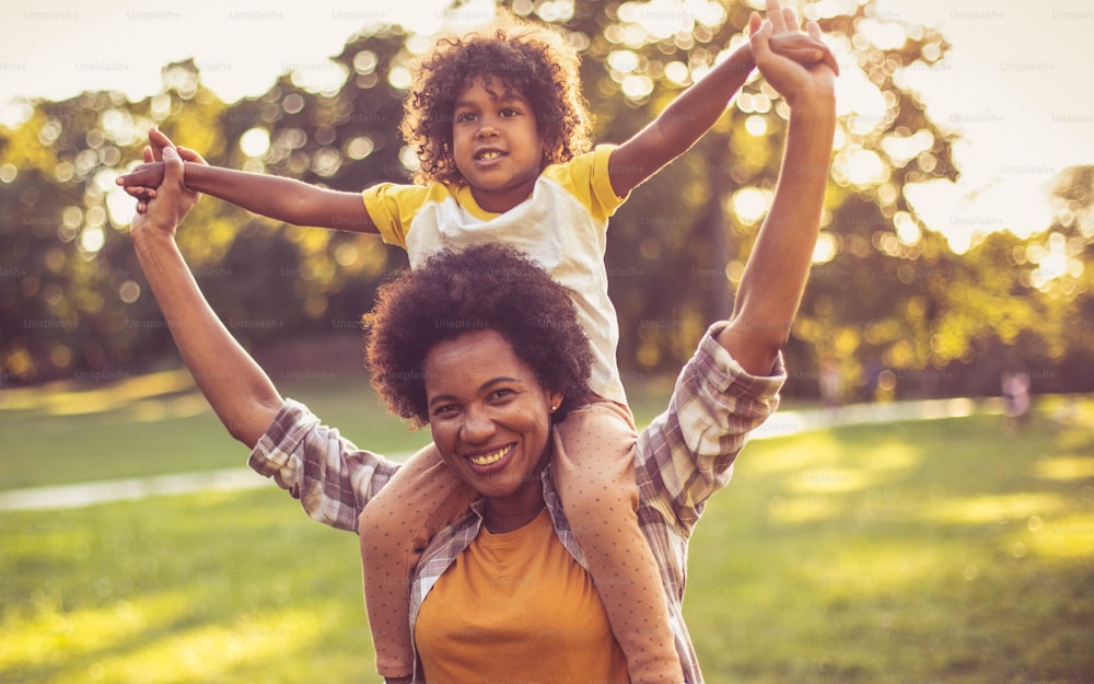 African American mother and daughter standing in park. Mother carrying her daughter on shoulders.