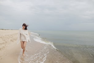 Bella donna spensierata con i capelli ventosi che corre sulla spiaggia sabbiosa alle onde fredde del mare, divertendosi. Giovane donna felice alla moda in maglione lavorato a maglia che si rilassa e si gode la vacanza sulla costa