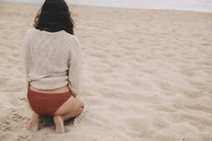 Hermosa mujer con cabello ventoso sentada en la playa de arena en el fondo del mar, momento despreocupado. Mujer joven con estilo en suéter de punto y bikini disfrutando de vacaciones y relajándose. Vista trasera