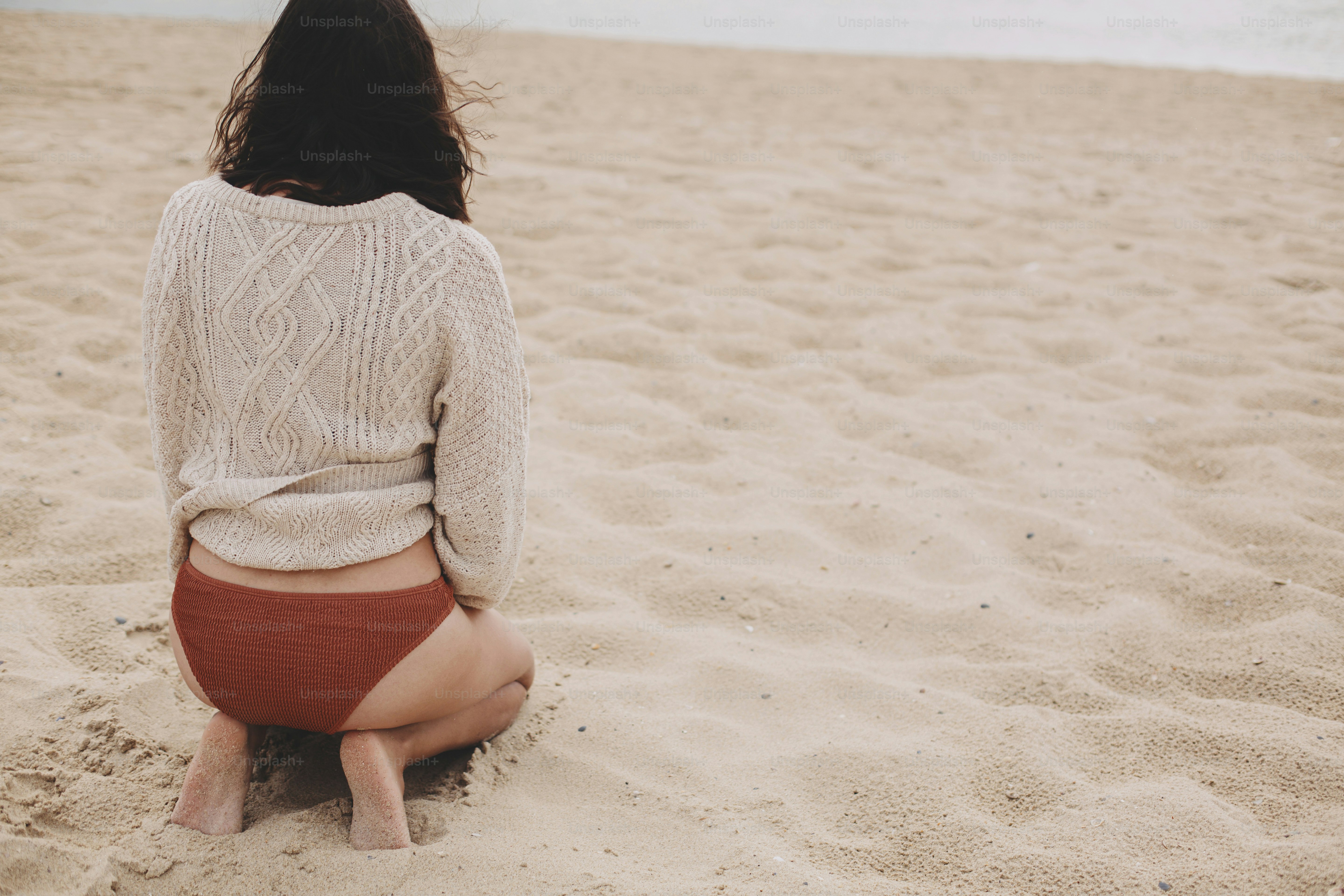 Beautiful woman with windy hair sitting on sandy beach on background of sea, carefree moment. Stylish young female in knitted sweater and bikini enjoying vacation and relaxing. Back view