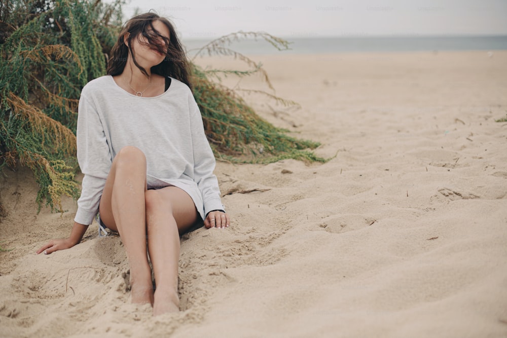 Mulher despreocupada bonita com cabelo ventoso e em suéter sentado na praia de areia no fundo da grama verde e do mar, momento tranquilo. Jovem fêmea elegante relaxando na costa. Clima de férias