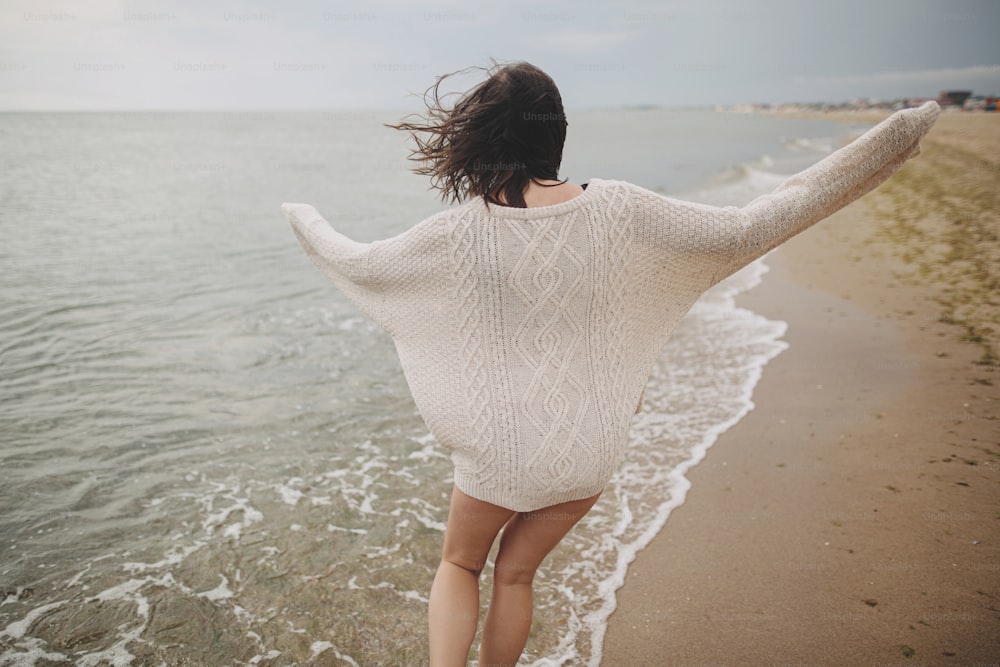 Mujer hermosa despreocupada en suéter de punto y con el cabello ventoso corriendo en la playa de arena en el mar frío, divirtiéndose. Mujer joven feliz con estilo que se relaja y disfruta de las vacaciones en la costa. Vista trasera