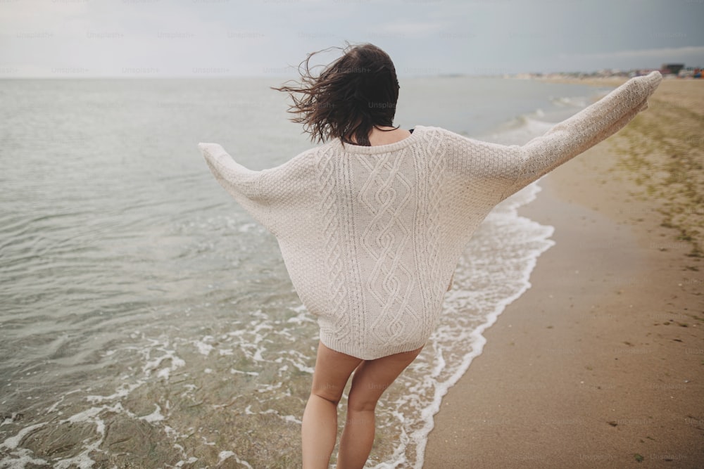 Carefree beautiful woman in knitted sweater and with windy hair running on sandy beach at cold sea, having fun. Stylish young happy female relaxing and enjoying vacation on coast. Back view