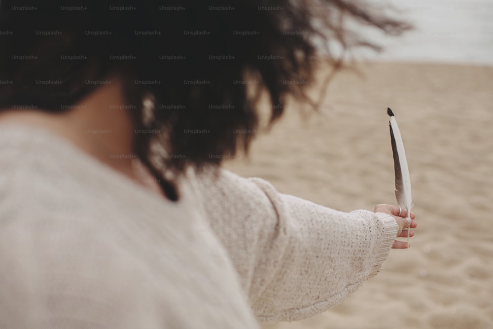 Feder in der Hand der schönen Frau mit windigen Haaren vor dem Hintergrund von Sandstrand und Meer, ruhiger Moment.  Frei und wild. Stilvolle junge Boho-Frau im Pullover mit Vogelfeder an der Küste.