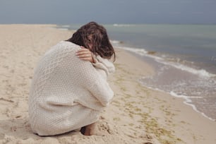 Carefree beautiful woman with windy curly hair sitting on sandy beach at sea, calm moment. Stylish young happy female in knitted sweater relaxing on coast. Summer vacation mood