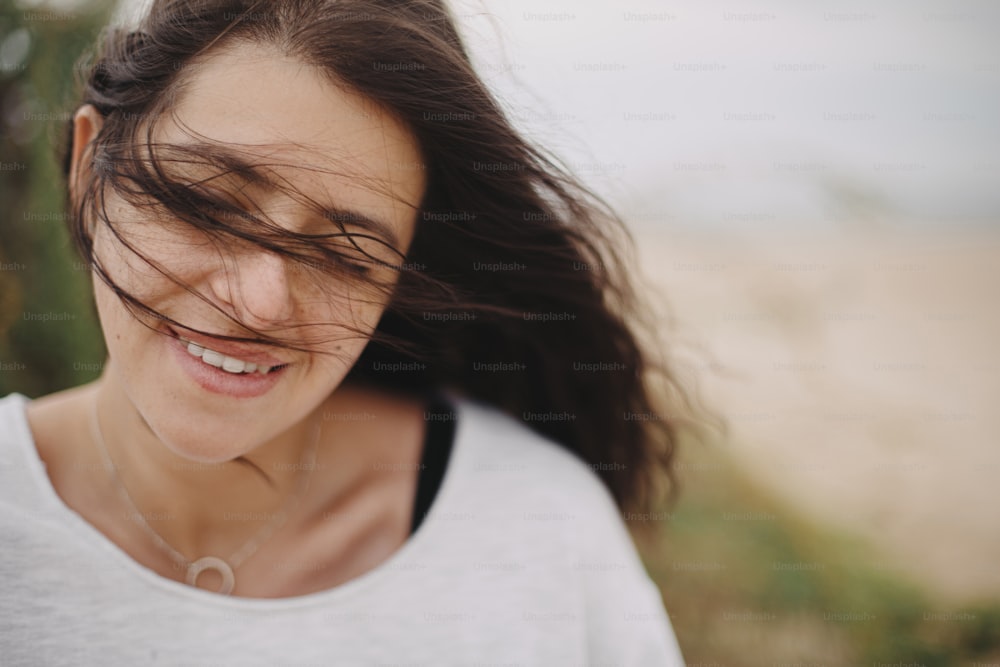 Beautiful happy woman with windy hair portrait on background of sandy beach and grass, carefree tranquil moment. Stylish young female in sweater relaxing and enjoying vacation on coast