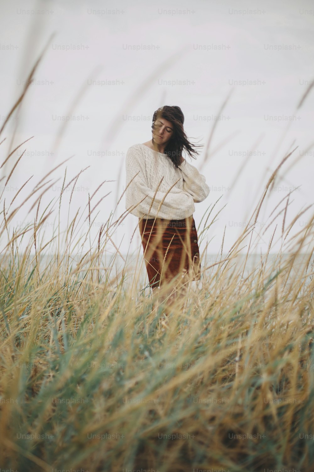 Beautiful stylish woman in knitted sweater and with windy hair posing among wild grass at sea. Authentic carefree moment. Fashionable young female standing on windy coast. Tranquility. Vertical