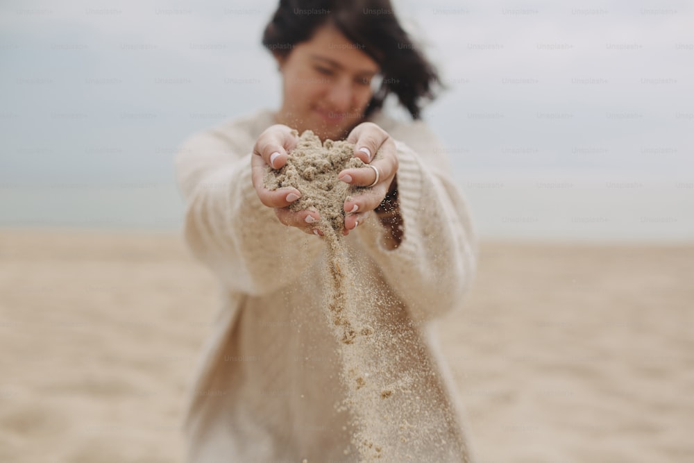 Stylish young woman in sweater releasing sand on beach, hands close up. Sand running through hands. Time concept. Summer beach and vacation. Woman hands holding sand, carefree moment