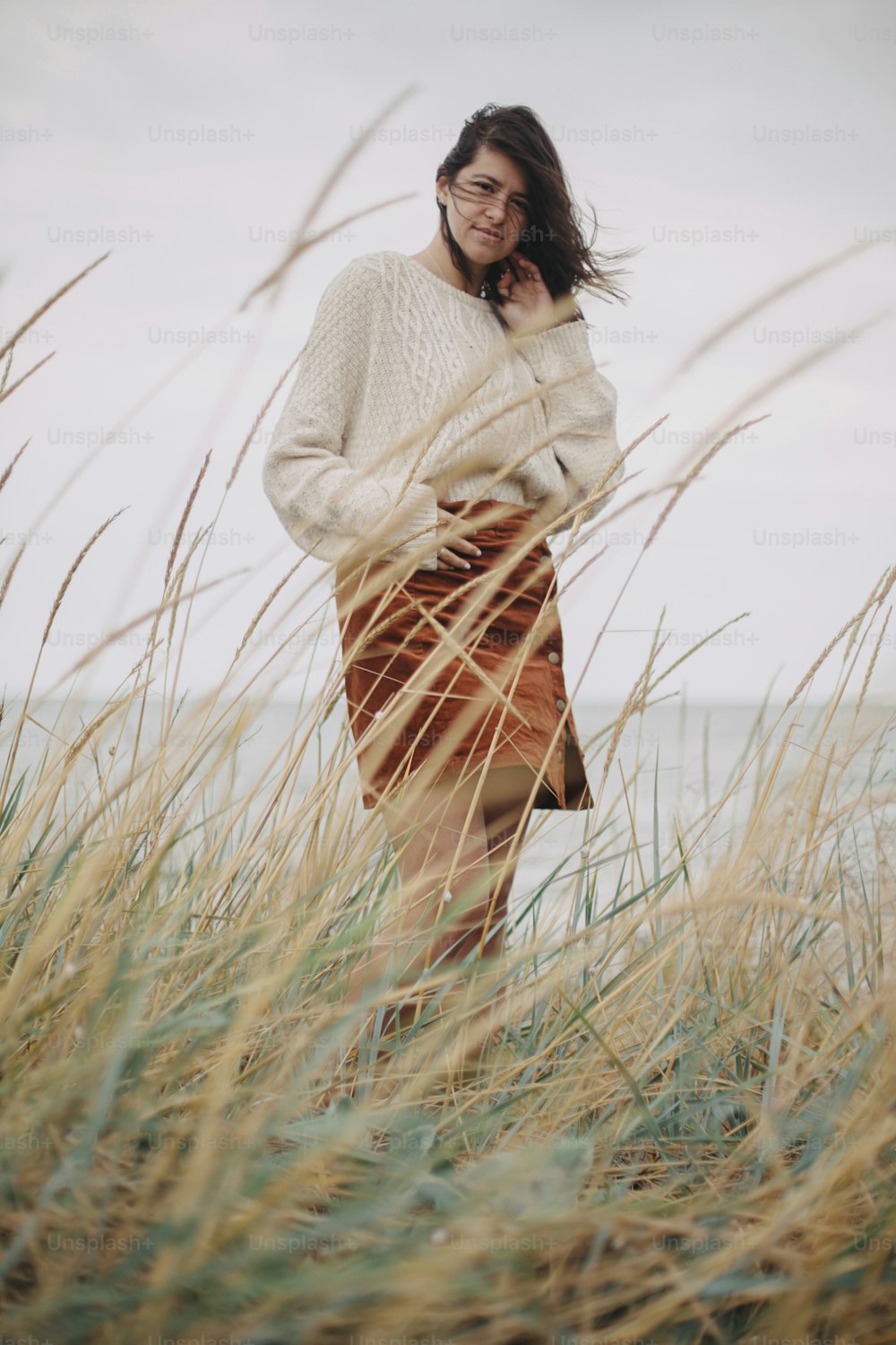 Schöne stilvolle Frau im Strickpullover und mit windigem Haar, die zwischen wildem Gras auf See posiert. Authentischer unbeschwerter Moment. Modische junge Frau, die an windiger Küste steht. Ruhe. Senkrecht