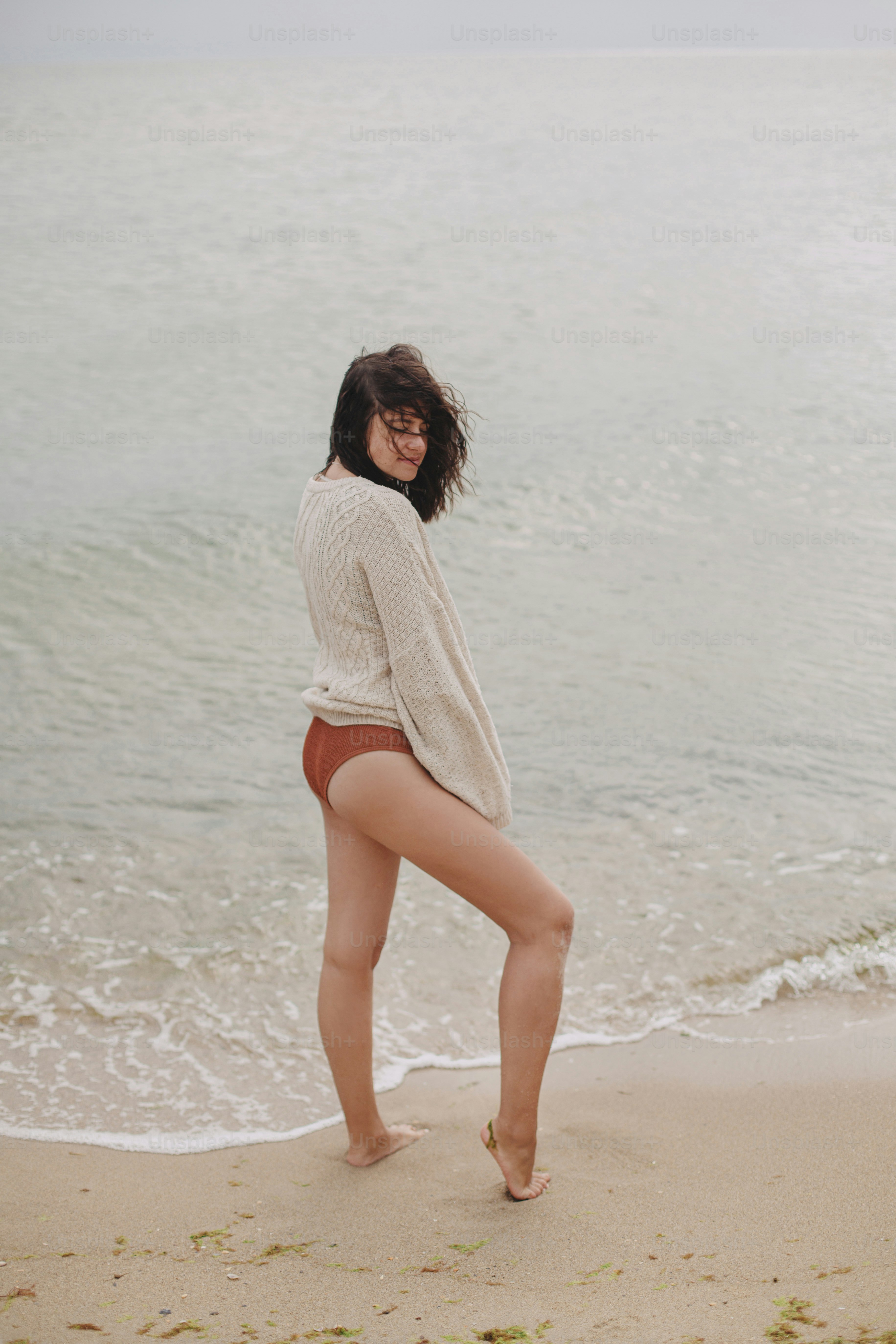 Carefree beautiful woman with windy hair in knitted sweater and bikini standing on sandy beach at sea. Stylish young sexy female posing and relaxing on coast. Tranquil moment, vacation mood