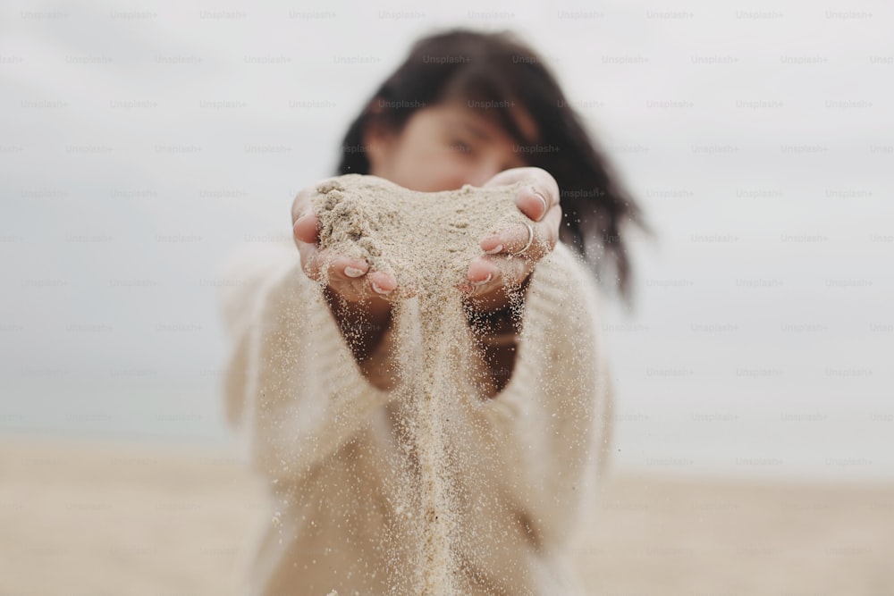 Woman hands holding sand on beach, carefree moment. Stylish young woman in sweater releasing sand on coast. Sand running through hands. Time concept. Summer beach and vacation