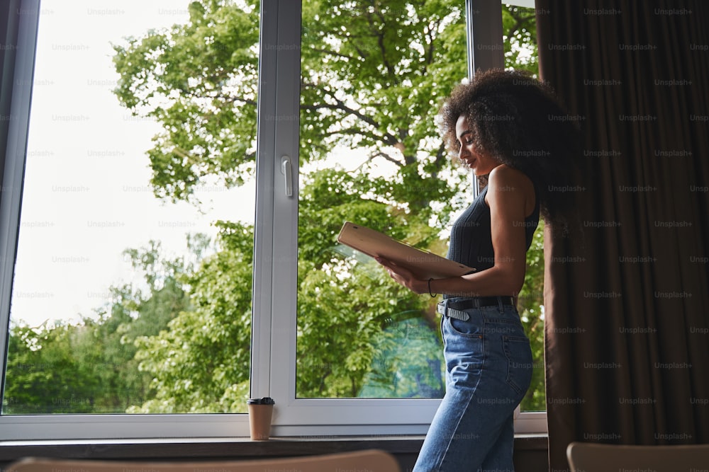 Smiling attractive lady in casual clothes looking at documents in a folder while standing alone. Coffee on the window