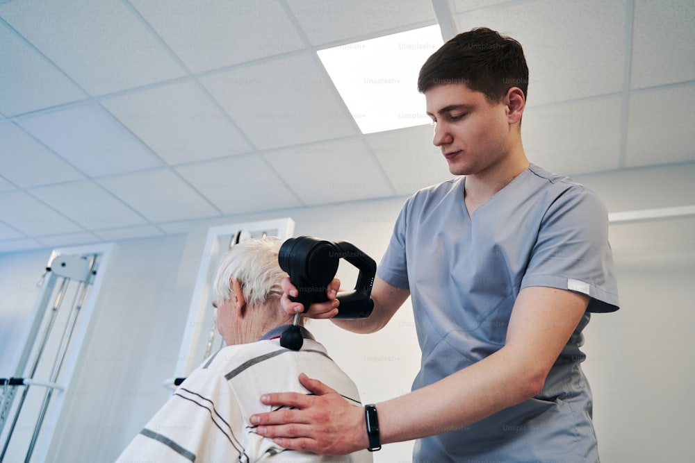 Caucasian physical therapist staying behind retired person and giving neck and upper back treatment with muscle massage gun