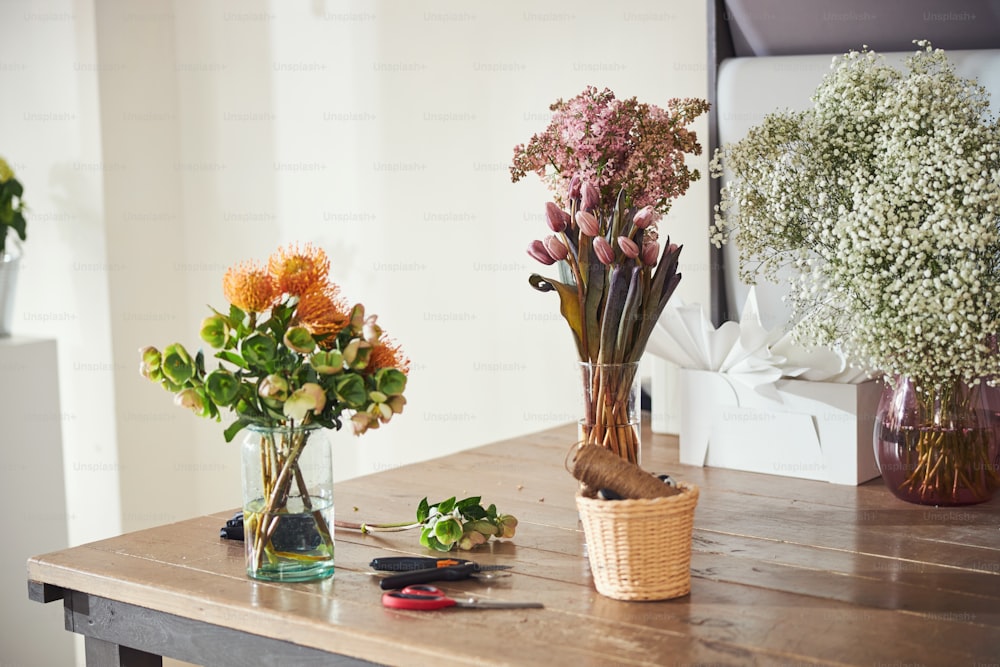 Three beautiful multi-colored bunches of fresh-cut flowers in clear glass vases filled with water placed on a sunlit wooden table