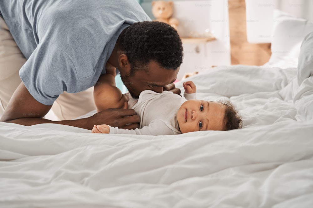 Love you so much. Portrait view of the brunette multiracial father kissing his cute baby, while little child resting on the bed at the bedroom. Fatherhood and happy childhood concept