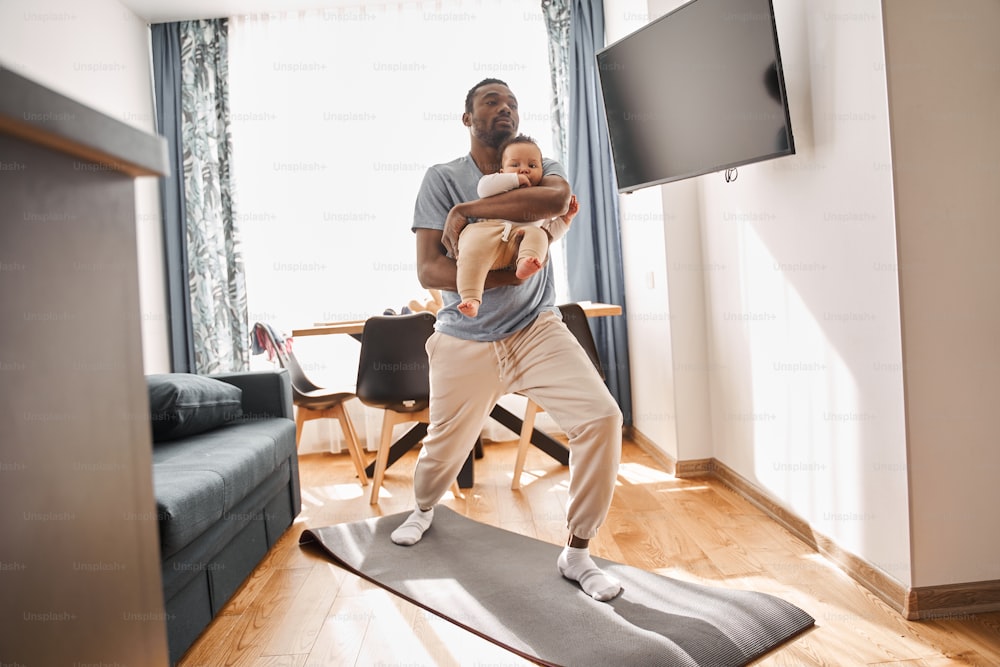 Peaceful focused man wearing domestic clothes standing at the mat with his son at his hands, while practicing yoga at his flat. No stress concept