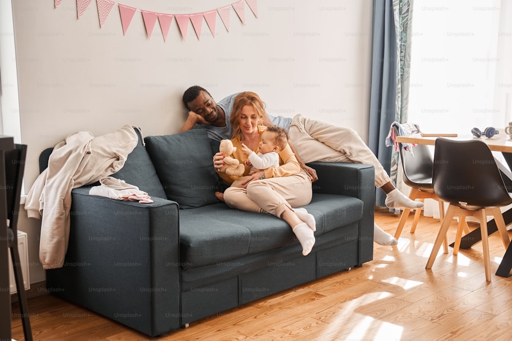 My dear. Full length view of the happy mother sitting at the sofa with her little newborn baby while spending time together with her husband at the bed at the morning. Stock photo