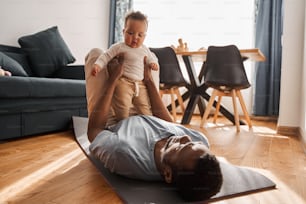 Comfortable posture. Full length view of the little brunette boy standing at the stomach of his father while he is exercising at the floor at home. Sport lifestyle with family concept