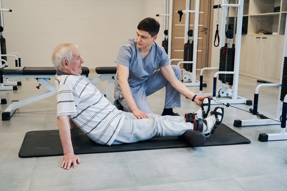 물리 치료사는 바닥에서 근육 재활 운동을 하는 동안 노인 다리를 가만히 잡고 있습니다.