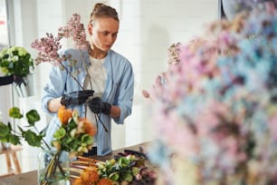 Serious blonde Caucasian female floral designer in casual clothes and nitrile gloves holding lilac branches